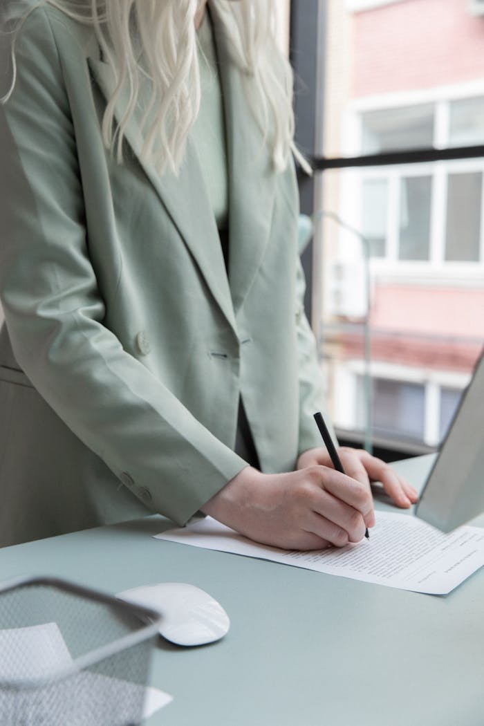 Crop faceless woman in formal clothes holding pen and writing notes on paper while working at table with computer and mouse